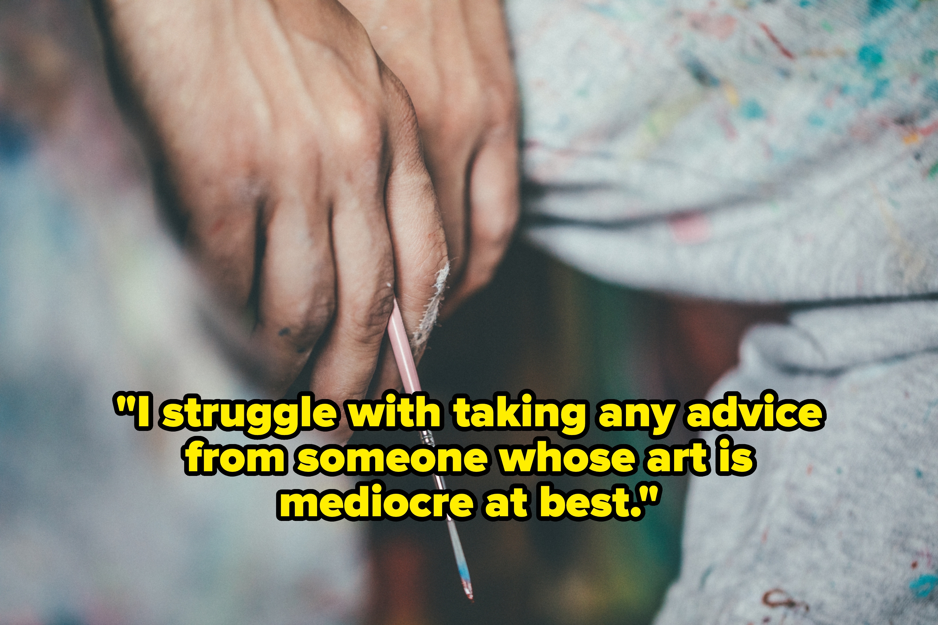 Hands holding a small paintbrush, resting on paint-splattered clothing, suggesting a creative and intimate setting