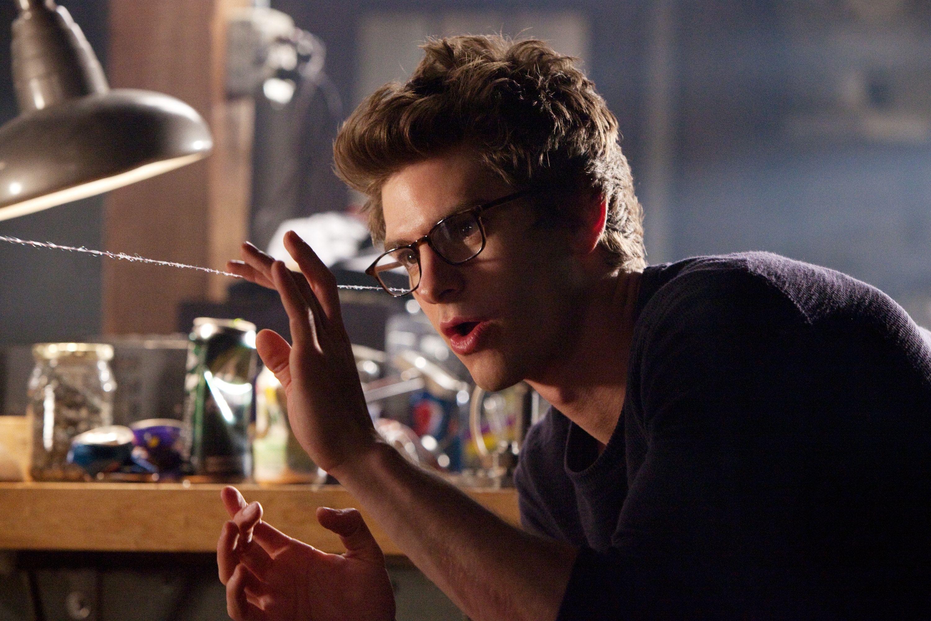 Person wearing glasses, gesturing with hand, in a workshop scene. Background includes tools and jars, creating a focused, contemplative atmosphere