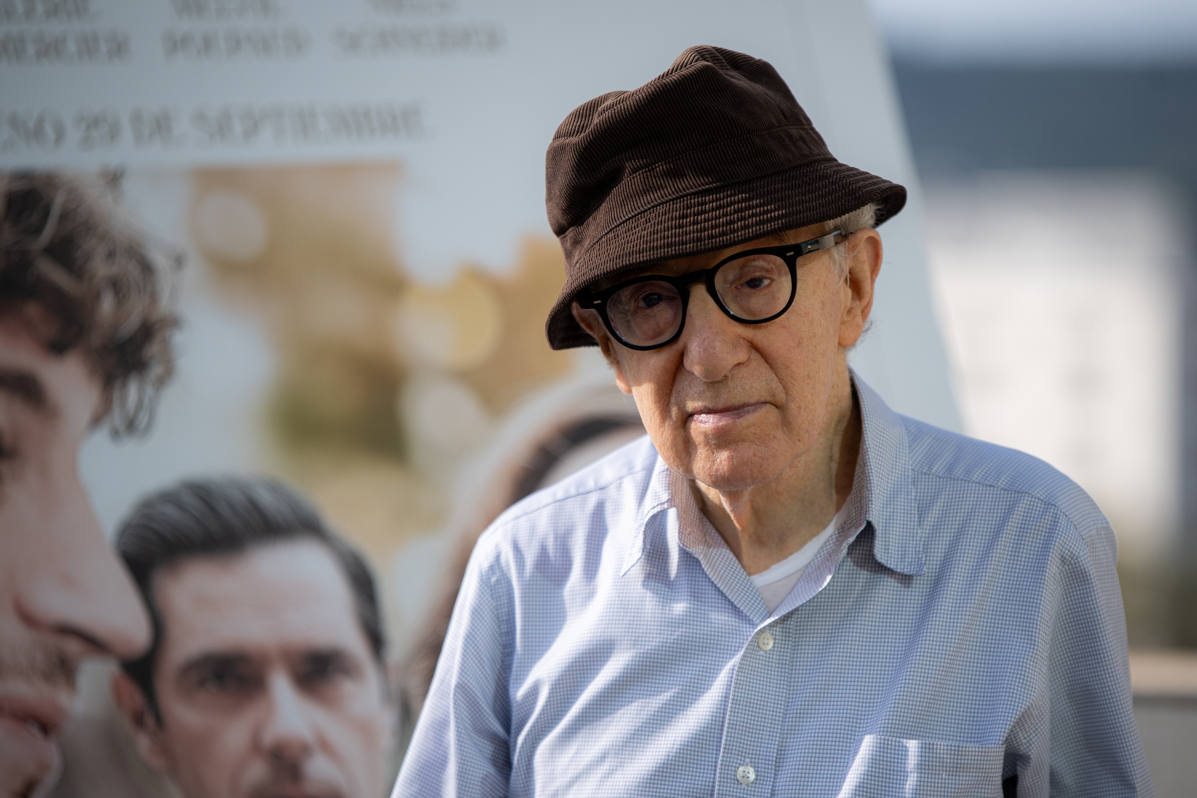 An elderly man wearing a hat and glasses stands in front of a movie poster or billboard, gazing forward