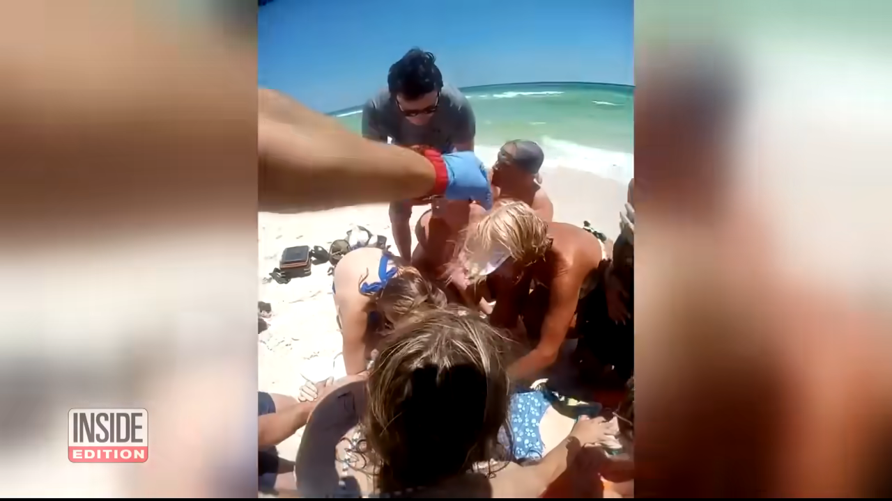 A group of people gather around someone on a beach, providing assistance. Waves are visible in the background