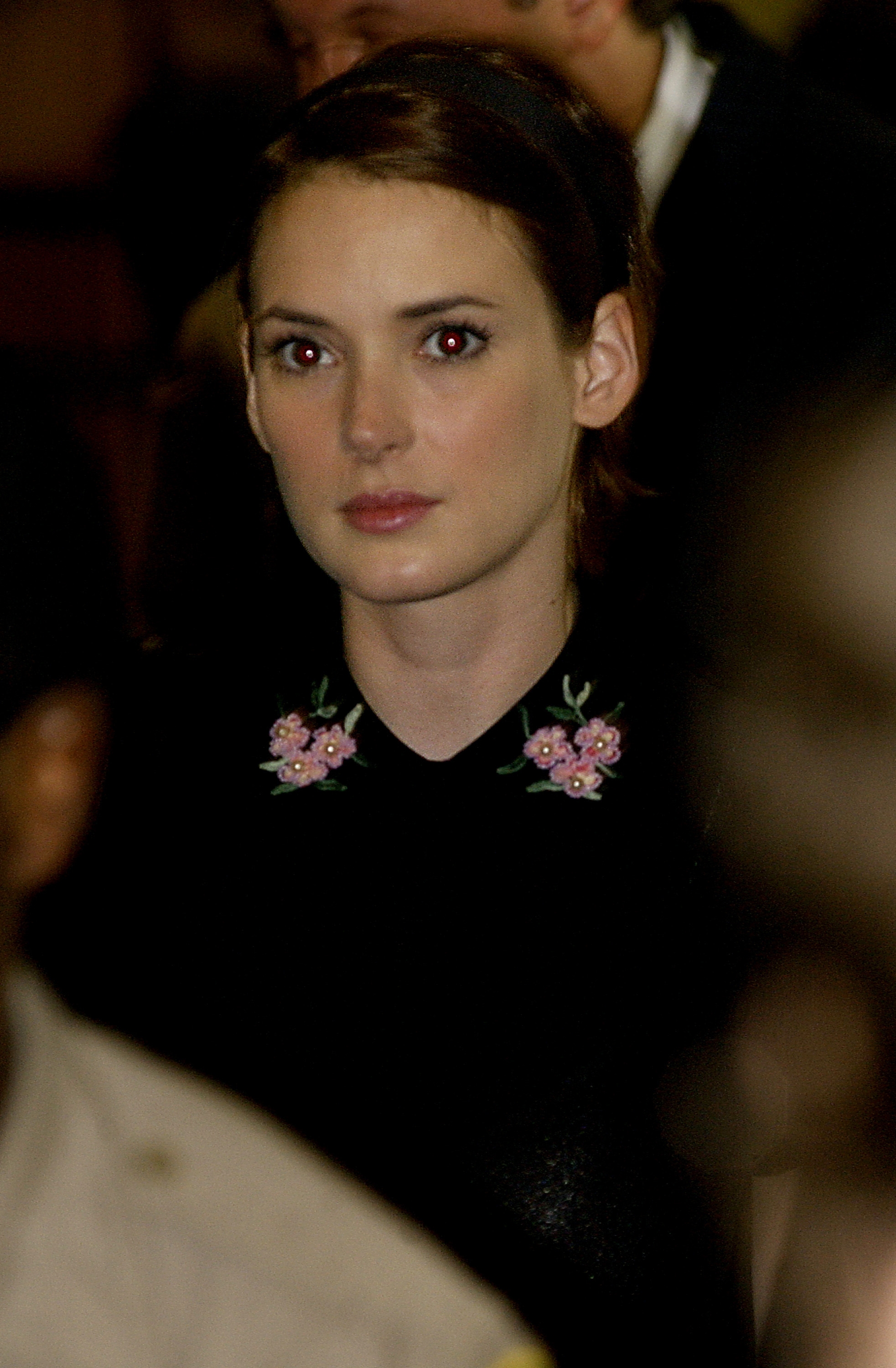 Person wearing a black top with floral embroidery on the collar, looking ahead, in a crowd