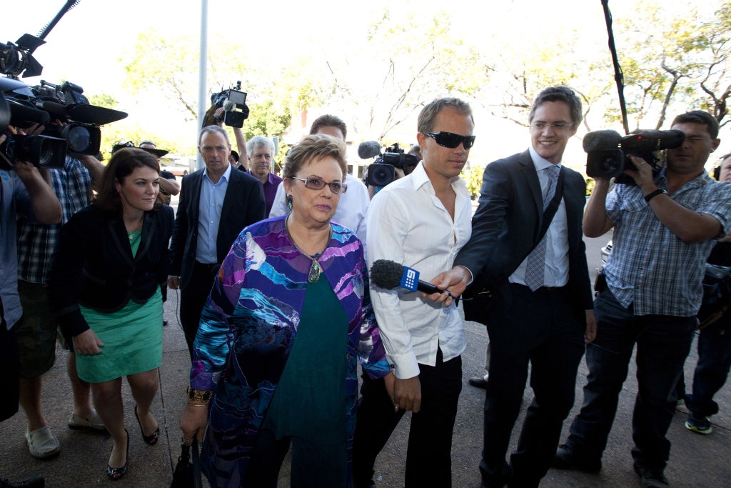 A group of photographers and journalists surrounds a woman in patterned clothing and a man in a white shirt with sunglasses