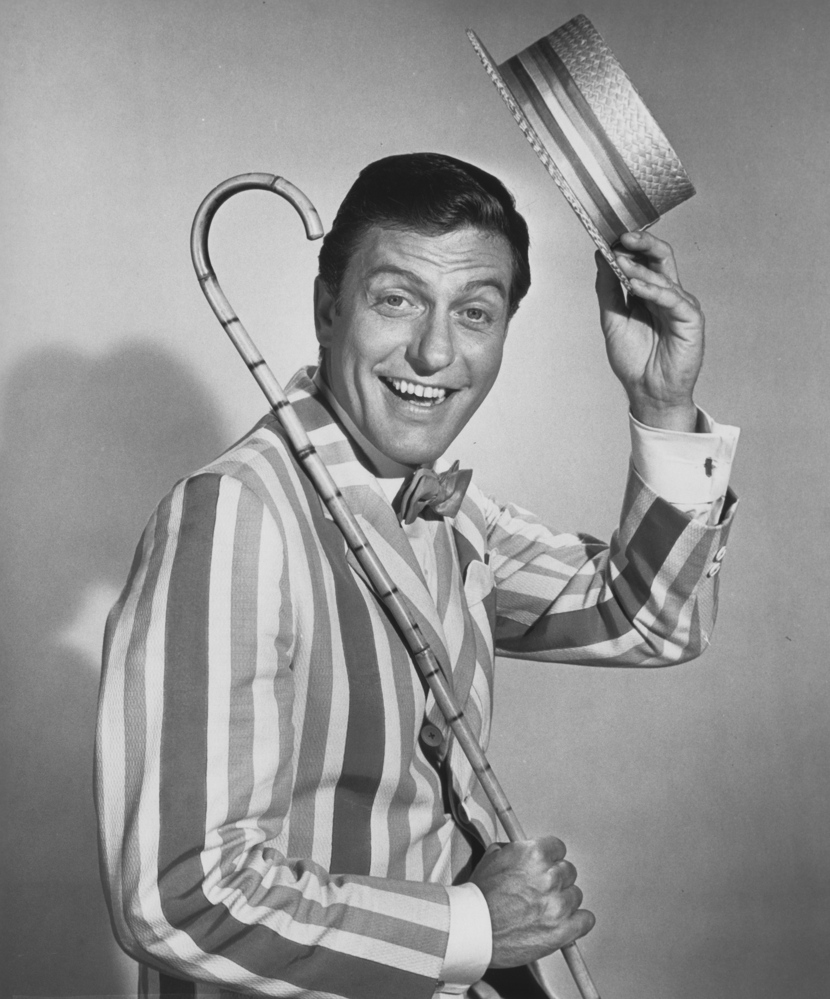 Person in striped jacket and bow tie, holding a cane and tipping a straw hat, smiling at the camera. Retro performance style