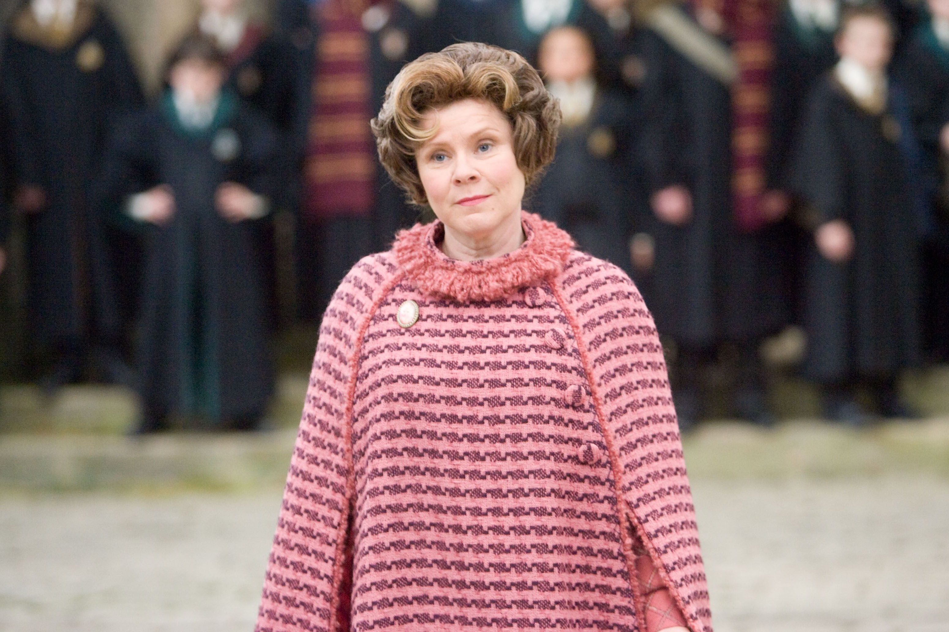 A character from a film wearing a patterned, high-collared poncho, standing in front of people in formal robes