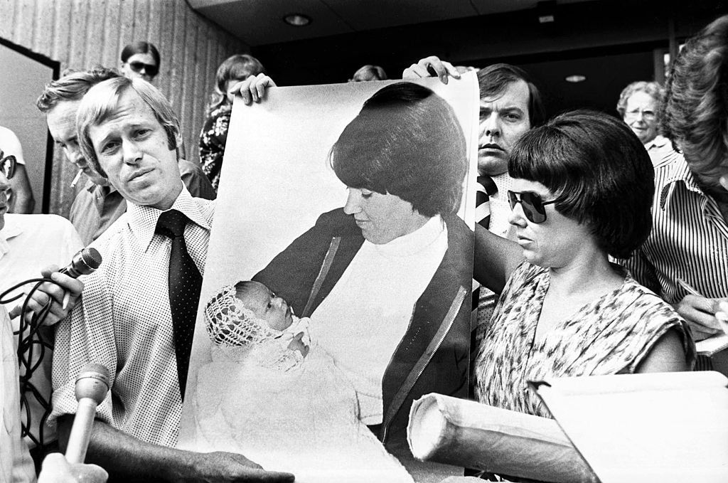 People holding a large photo of a woman with a baby, surrounded by reporters and microphones in a public setting