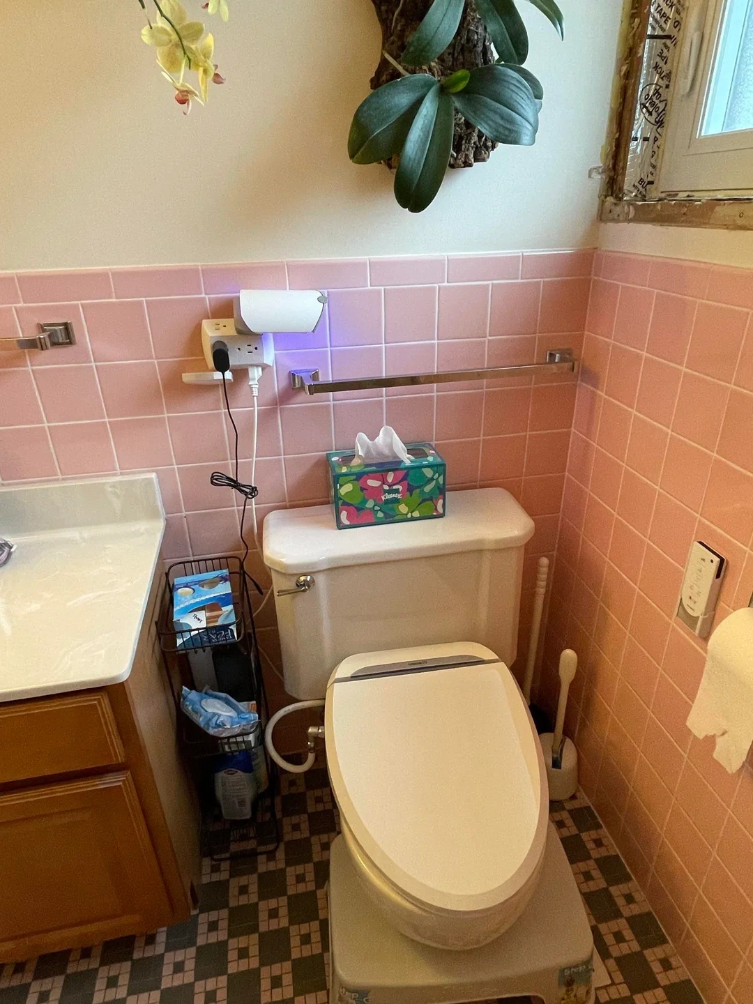 A bathroom with a pink-tiled wall, featuring a toilet, a tissue box on top, and cleaning supplies nearby
