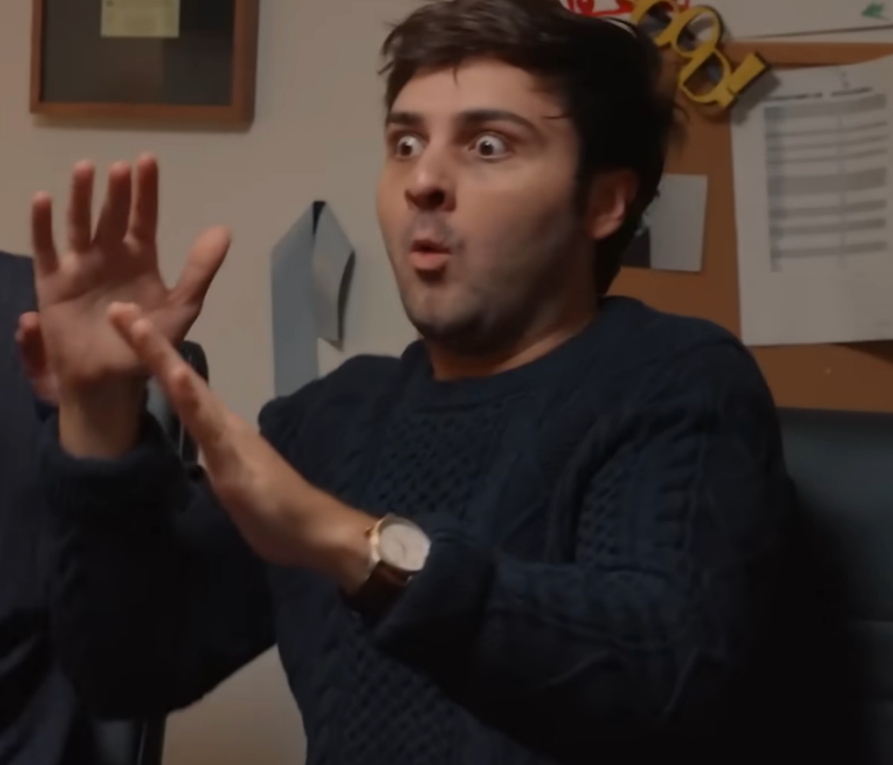 Person with a surprised expression gestures with hands, wearing a knit sweater and watch, seated in an office setting
