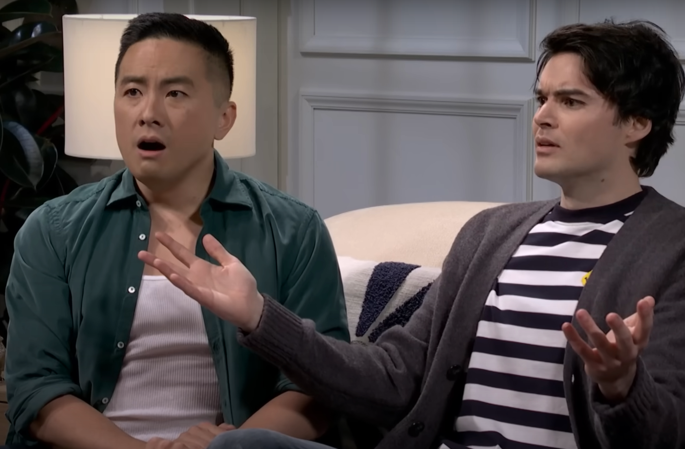 Two people sitting on a couch, looking surprised and confused, gesturing with open hands. One wears a cardigan over a striped shirt, the other a shirt over a tank top