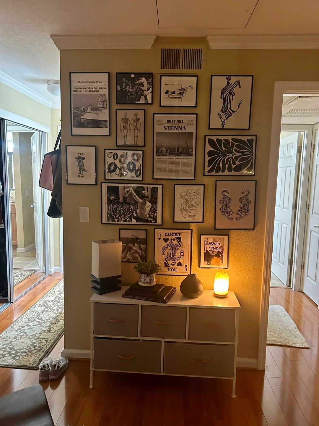 Gallery wall with various framed art and photos above a small dresser, softly lit by a lamp. Cozy home atmosphere with decor items