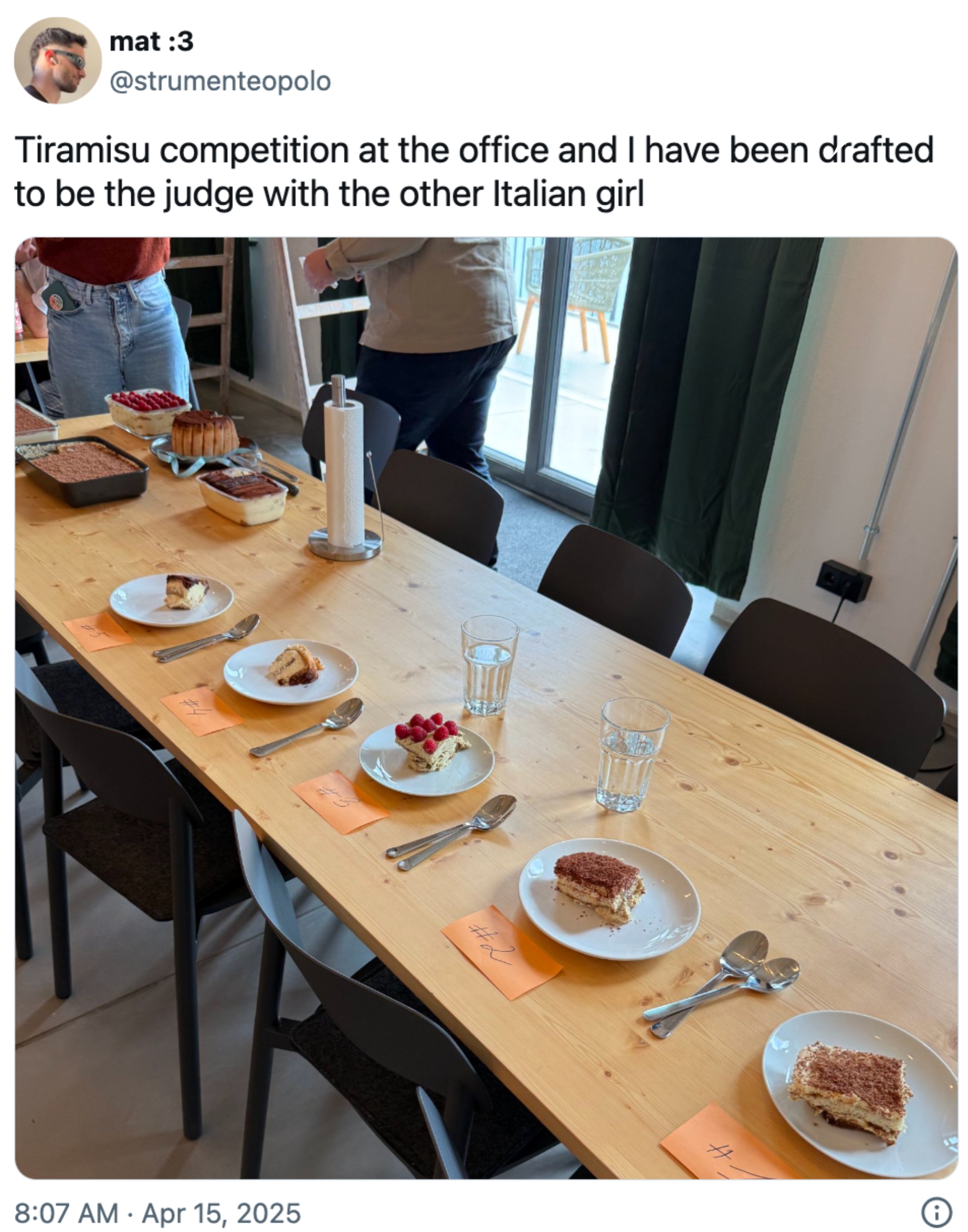 Table setup for a tiramisu competition with several plated slices labeled with orange notes, and a person standing in the background