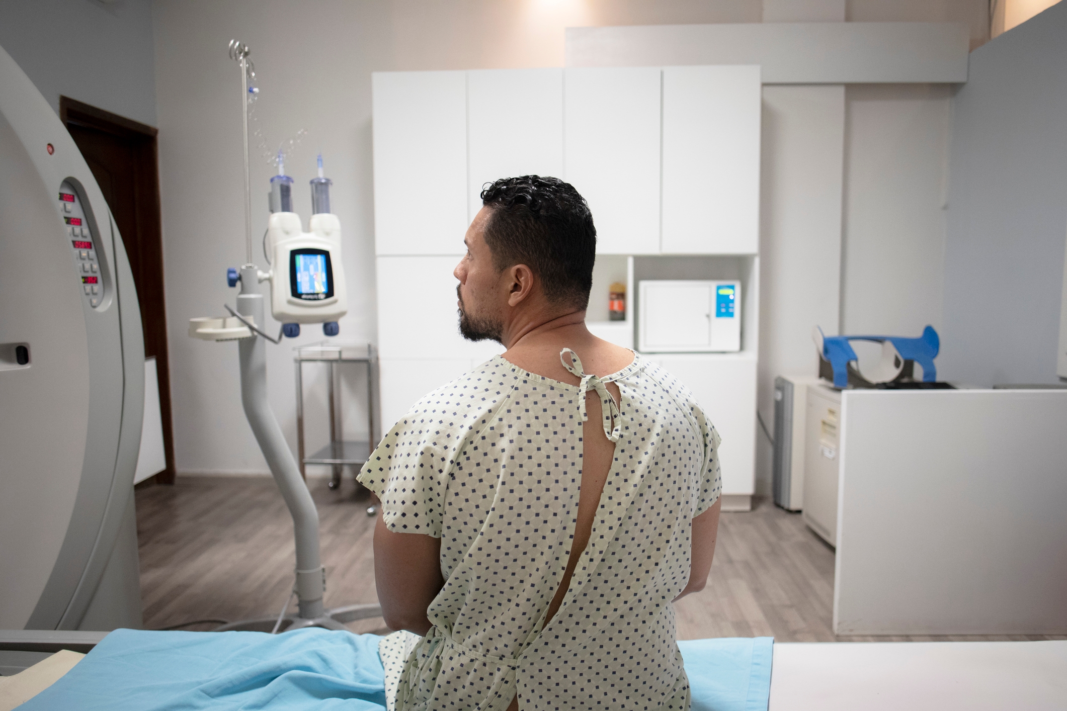 A patient in a hospital gown sits on a bed next to medical equipment, likely awaiting a procedure in a clinical setting
