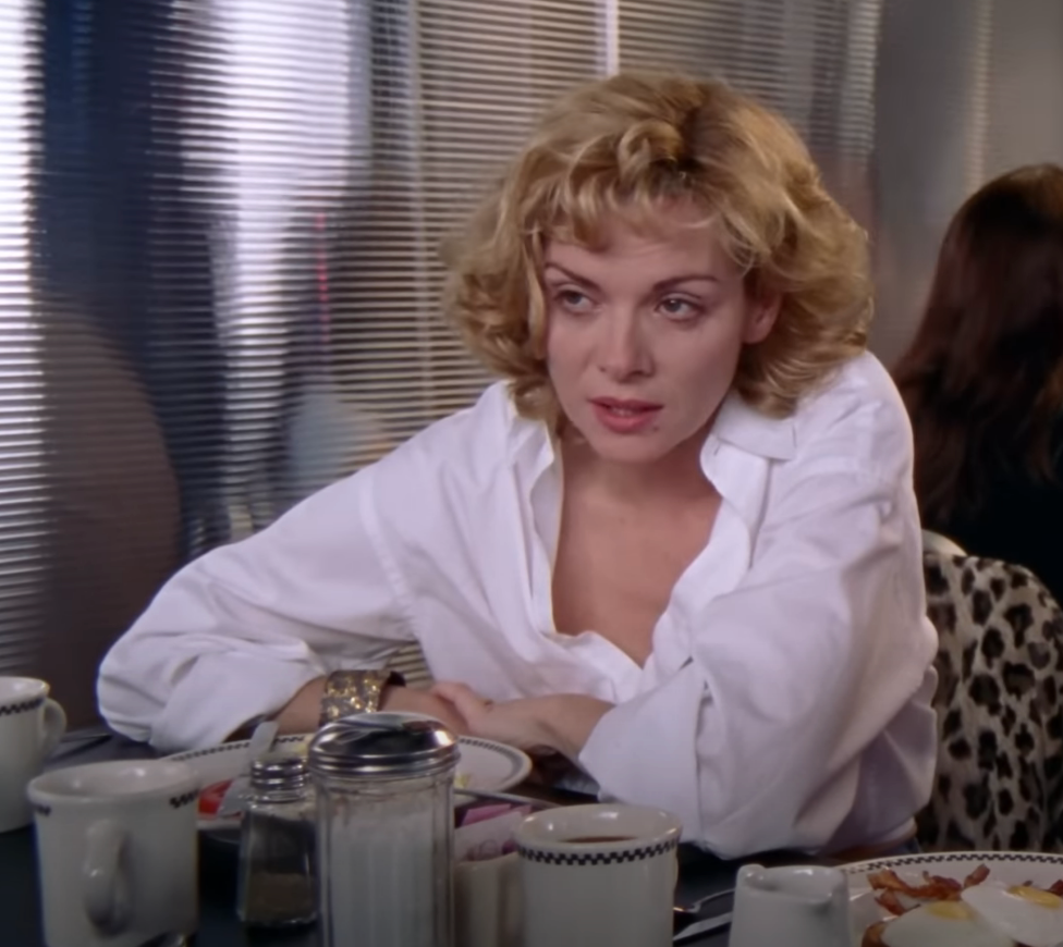 Person with short curly hair in a diner, wearing a white shirt, with a neutral expression, sitting at a table with plates and cups