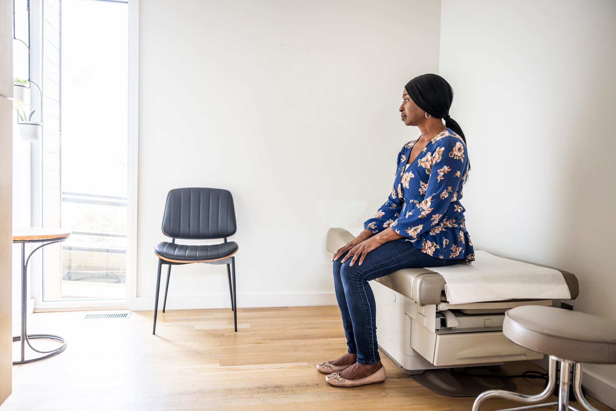 Person sits on an exam table in a doctor's office, facing sideways, wearing a floral top and jeans