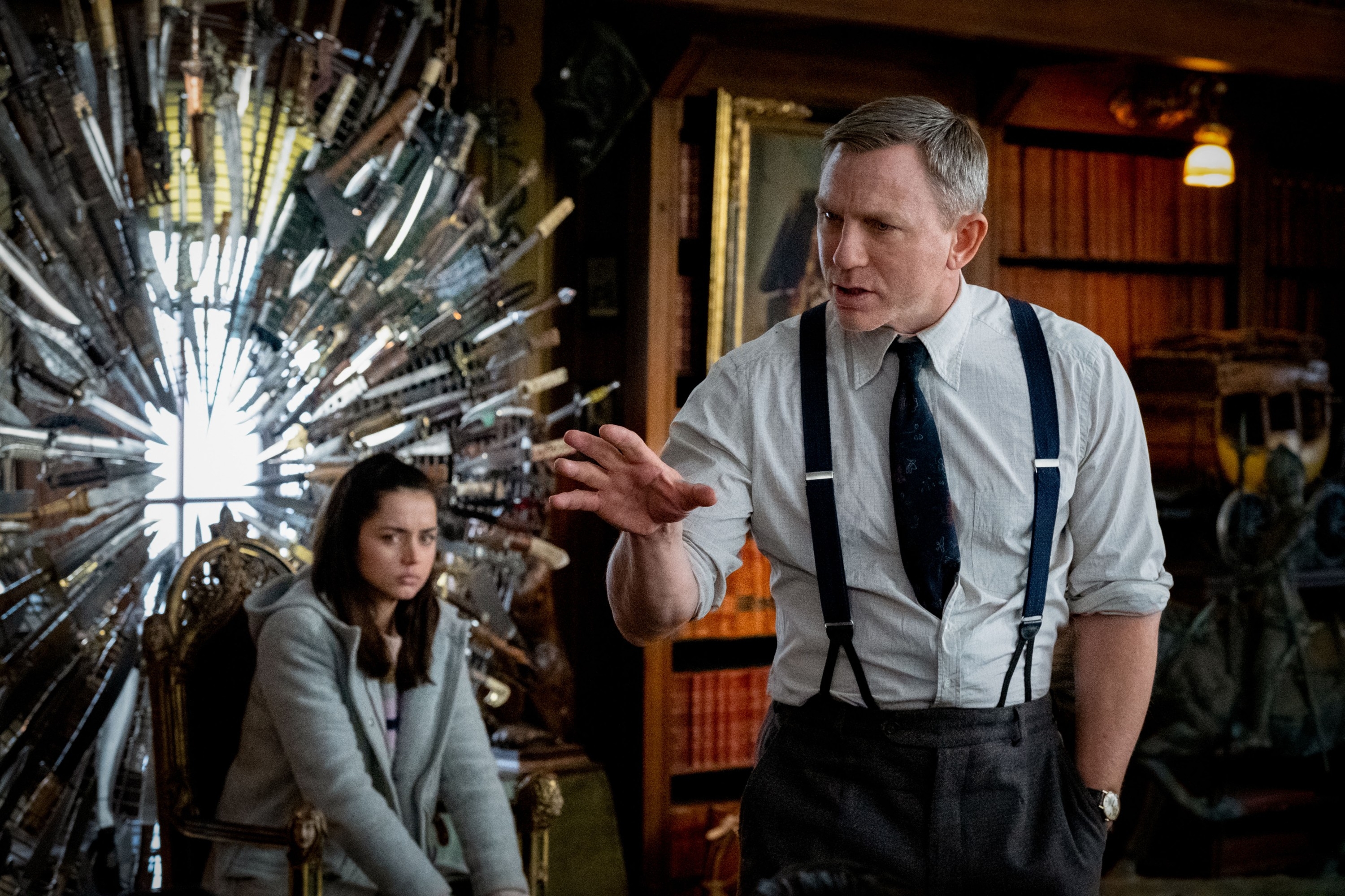 A detective in suspenders gestures while speaking to a woman seated near a knife-covered chair, in a scene from a movie set