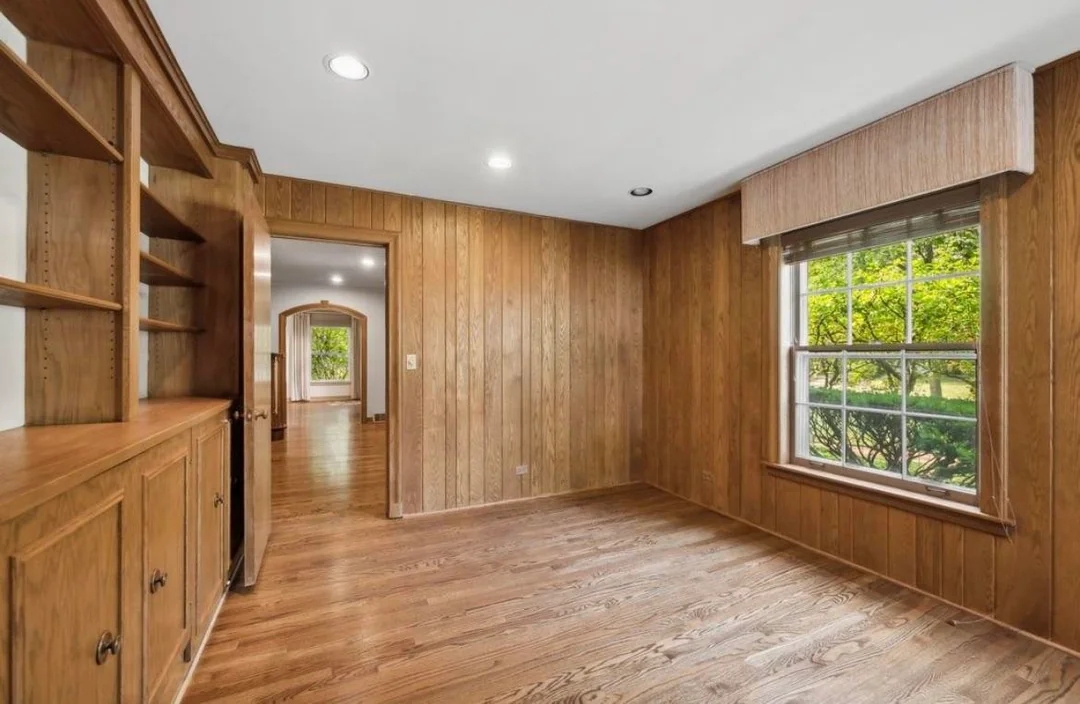Empty wooden-paneled room with built-in shelves, a large window, and an open doorway leading to another room with visible greenery outside