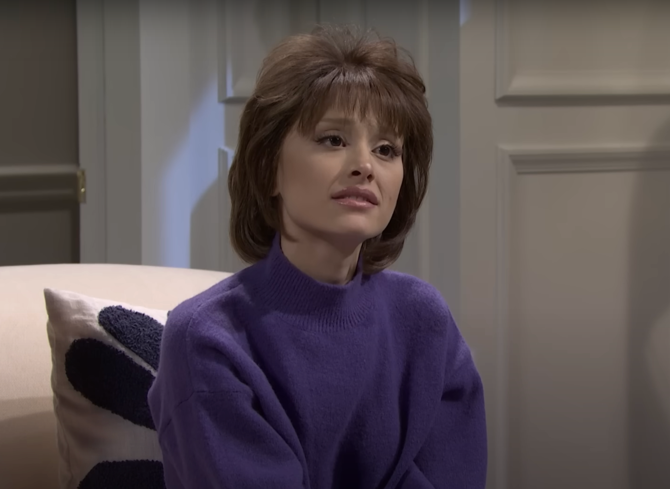 A person with short hair wearing a sweater is seated indoors, looking contemplative