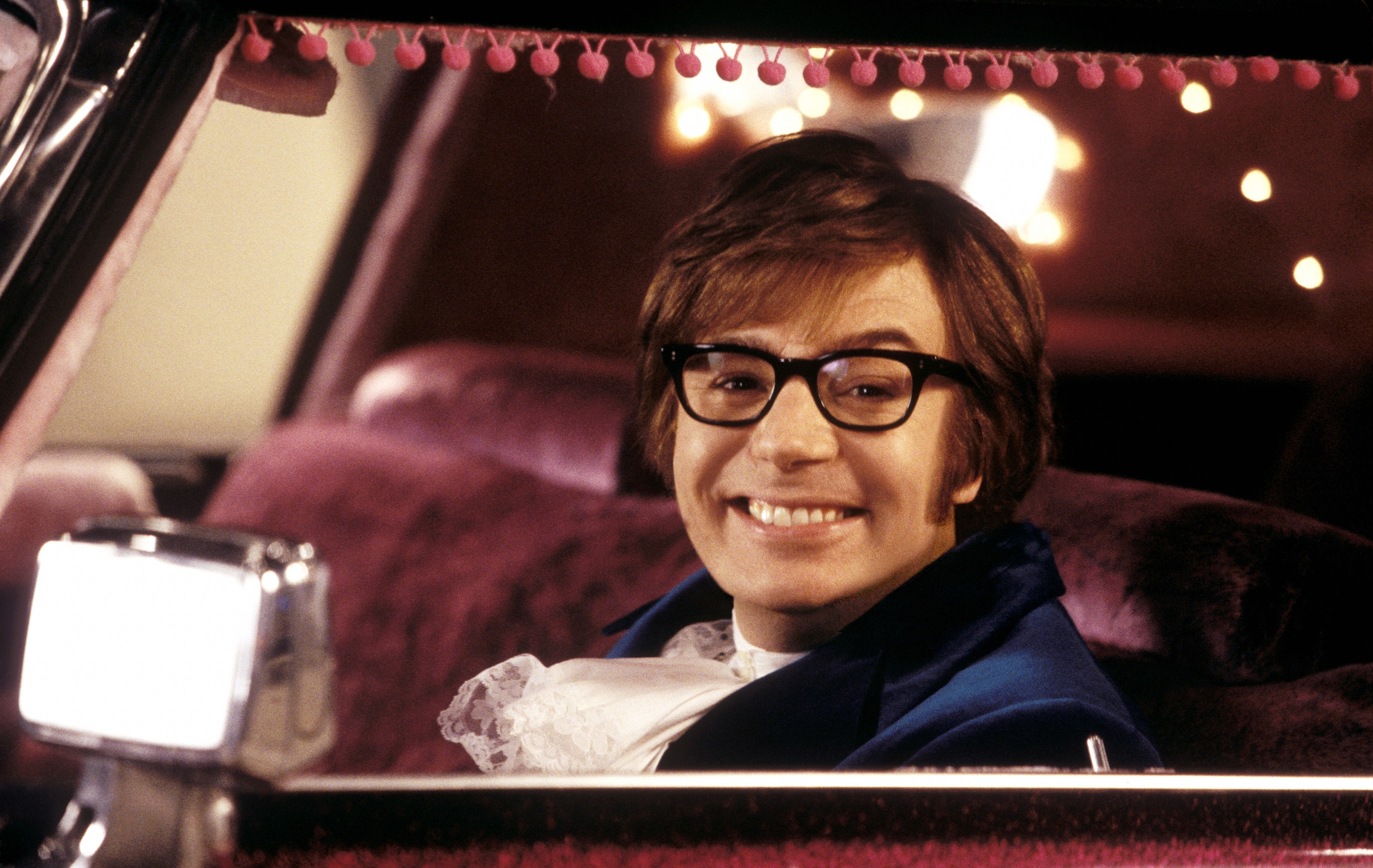 Person in shaggy 1960s-style suit and glasses smiling from car seat, with retro decor accents visible