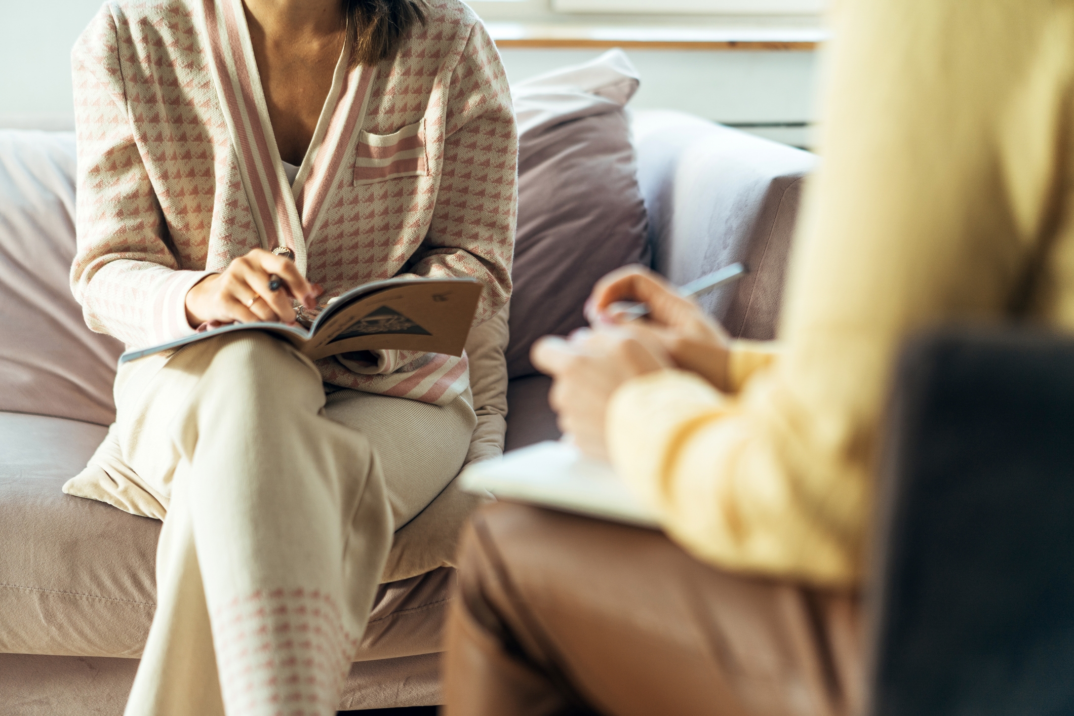 Two individuals sit on a couch, engaged in a discussion, each holding an open notebook and pen, suggesting a meeting or counseling session