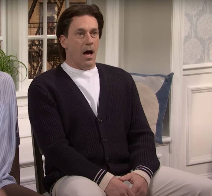 A person sitting indoors, wearing a cardigan and white shirt, appears surprised with their mouth open