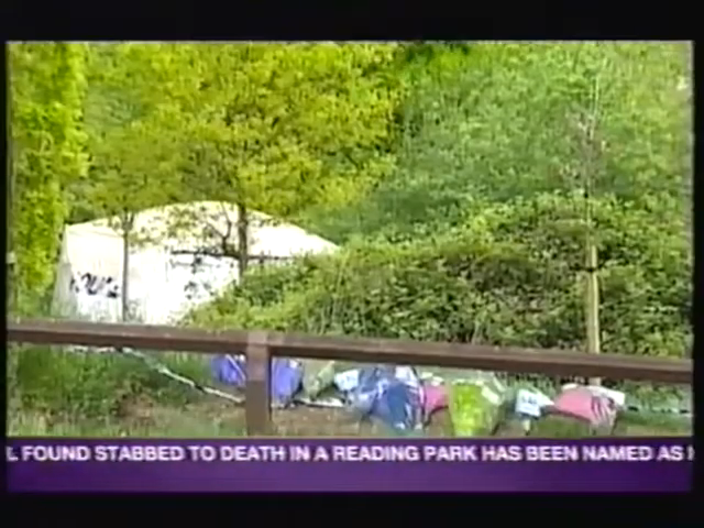 Tents are set up in a wooded area, likely a campsite. A wooden railing is in the foreground. News ticker text is partially visible at the bottom