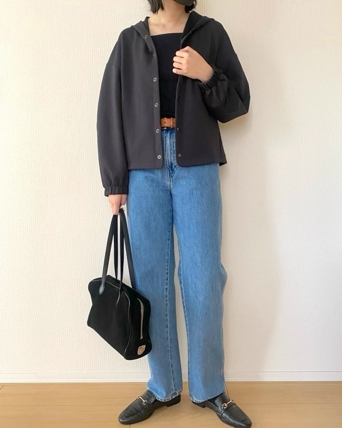 女性がカジュアルな服装でジーンズとジャケットを着て立っている。黒いバッグを持ち、リラックスした姿勢。