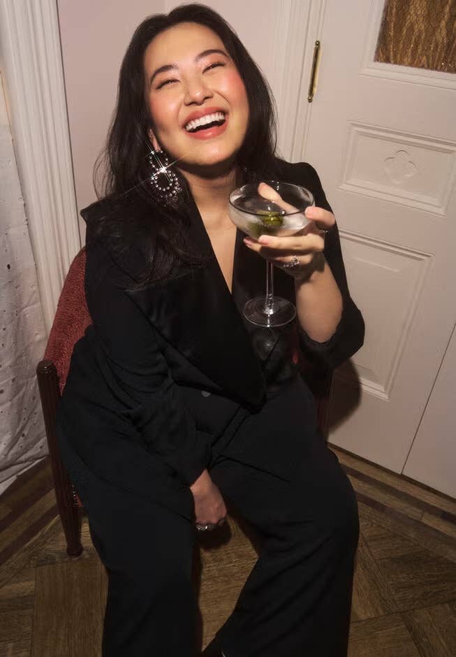 Person laughing, seated in elegant dark outfit, holding a martini glass with an olive