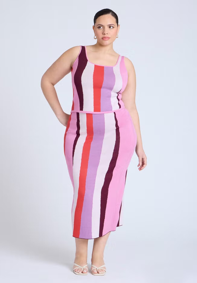 Model wears a form-fitting, striped tank top and matching skirt with heels, posing confidently against a plain backdrop