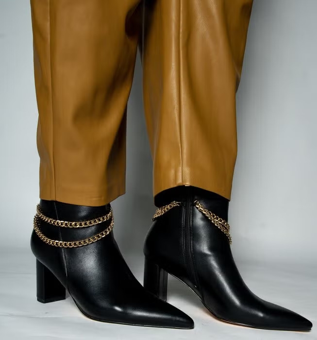 Black pointed ankle boots with gold chain details are styled with brown leather pants