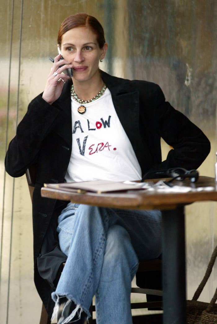 Julia Roberts sits at a café, talking on the phone. They wear a casual blazer and jeans, with a unique graphic tee and necklace