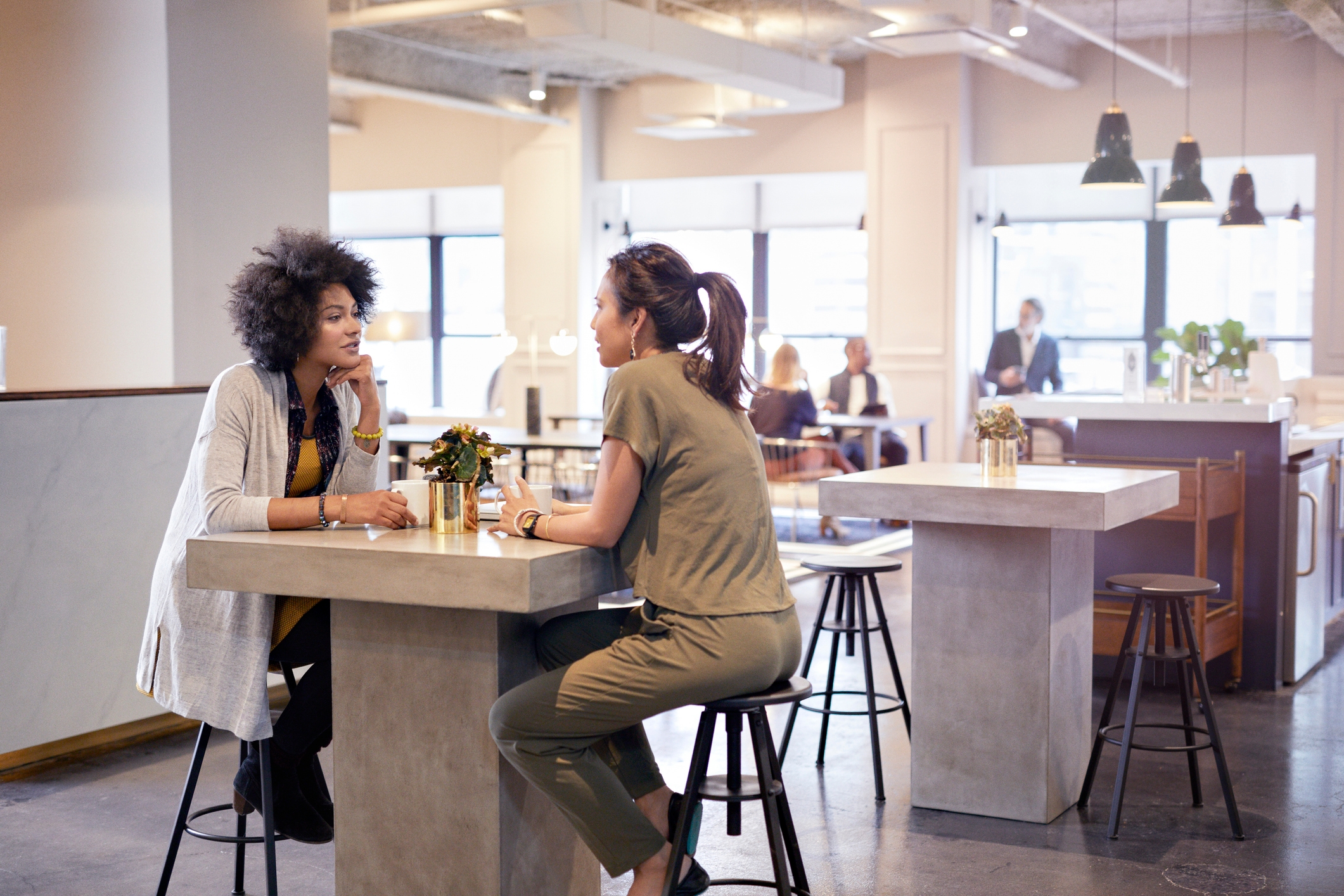 Two people sitting at a high-top table in a modern cafe-style setting, engaged in conversation. Other patrons are in the background