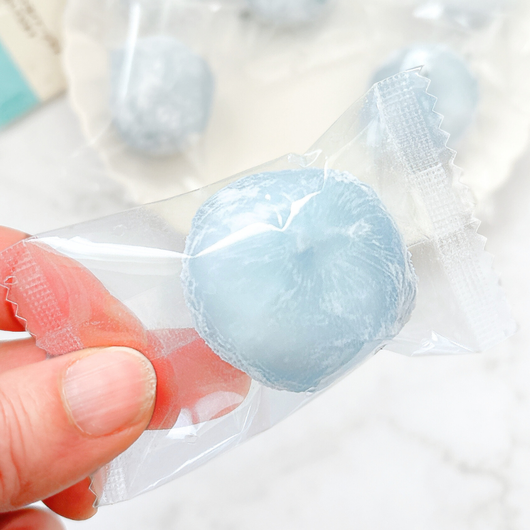 手に持たれた個別包装の青い大福。後ろには複数の大福がぼんやりと見えます。