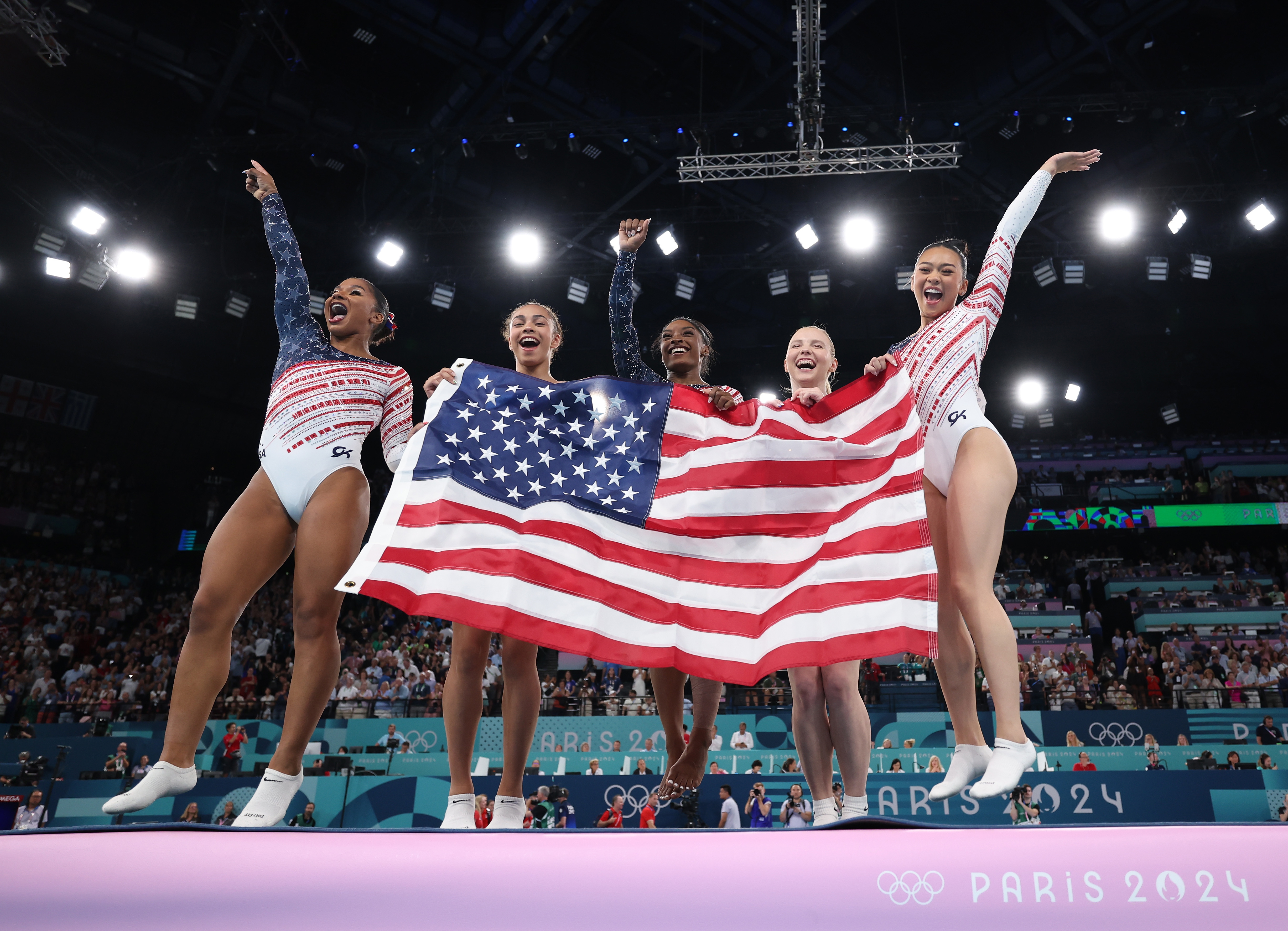 Five gymnasts in leotards hold a U.S. flag, celebrating their win at the Paris 2024 Olympics, smiling and cheering on the podium