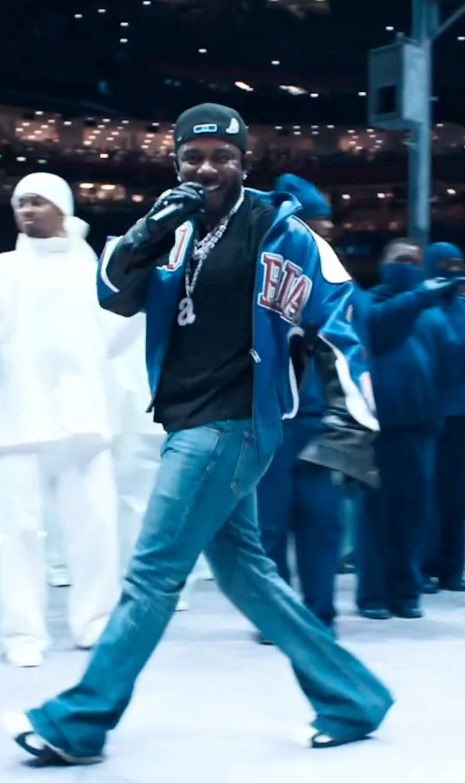 Kendrick Lamar energetically performs on stage, wearing a blue jacket, jeans, and a cap, with backup dancers in white and blue outfits in the background
