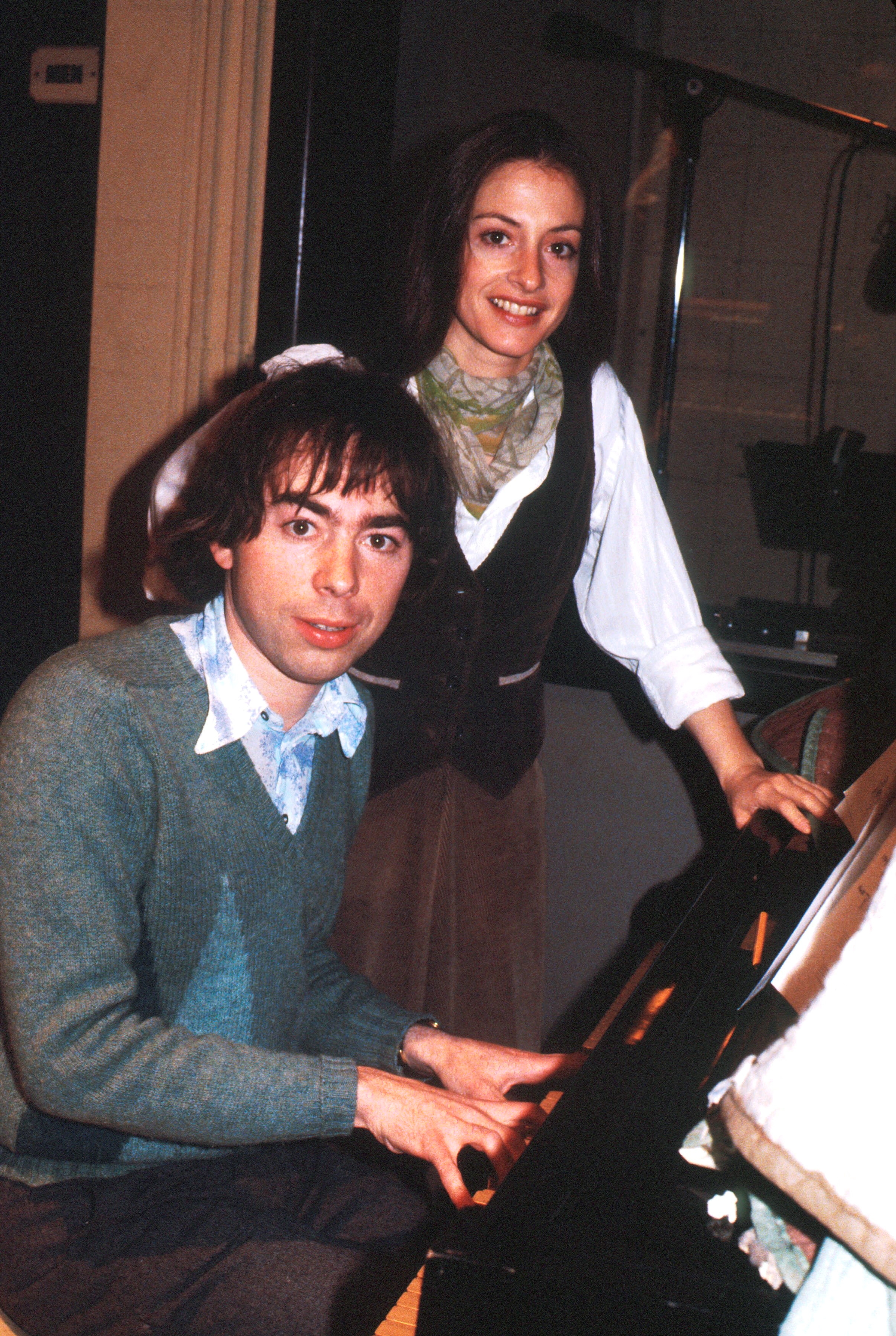 Two people in a music studio, one seated playing a piano, and the other standing beside, both casually dressed and smiling at the camera