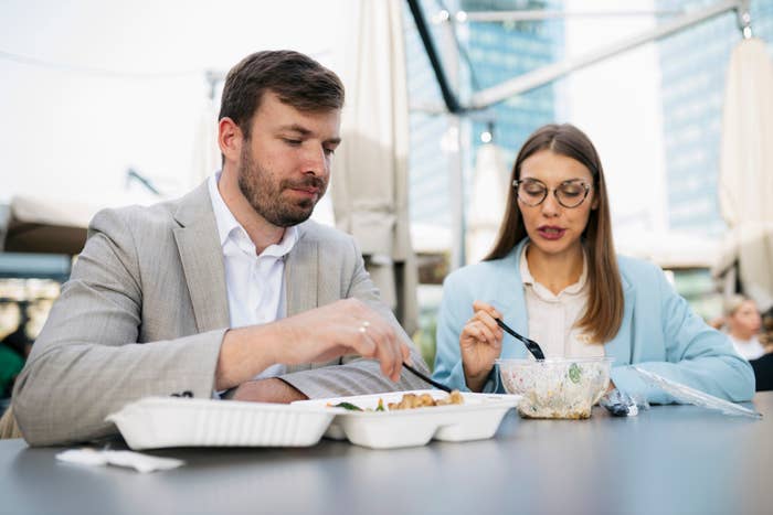 Two people in business attire eating from takeout containers at an outdoor table. Skyscrapers are visible in the background