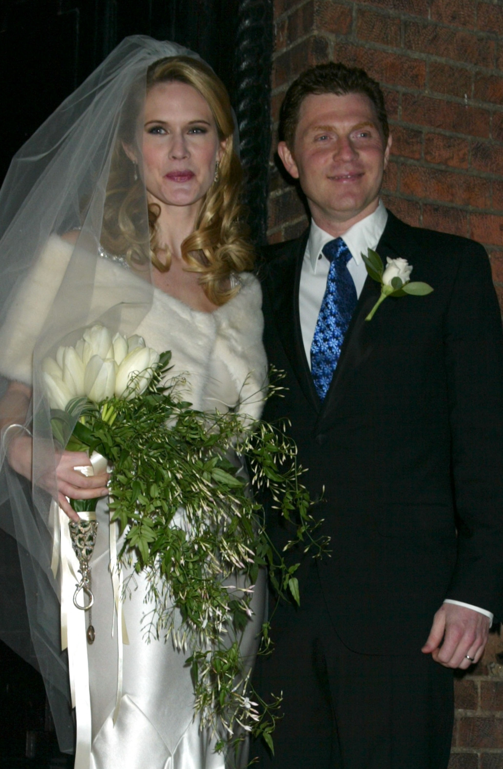 A couple in wedding attire poses together, smiling. The bride holds a bouquet with greenery, wearing a veil and satin dress. The groom wears a suit with a floral tie