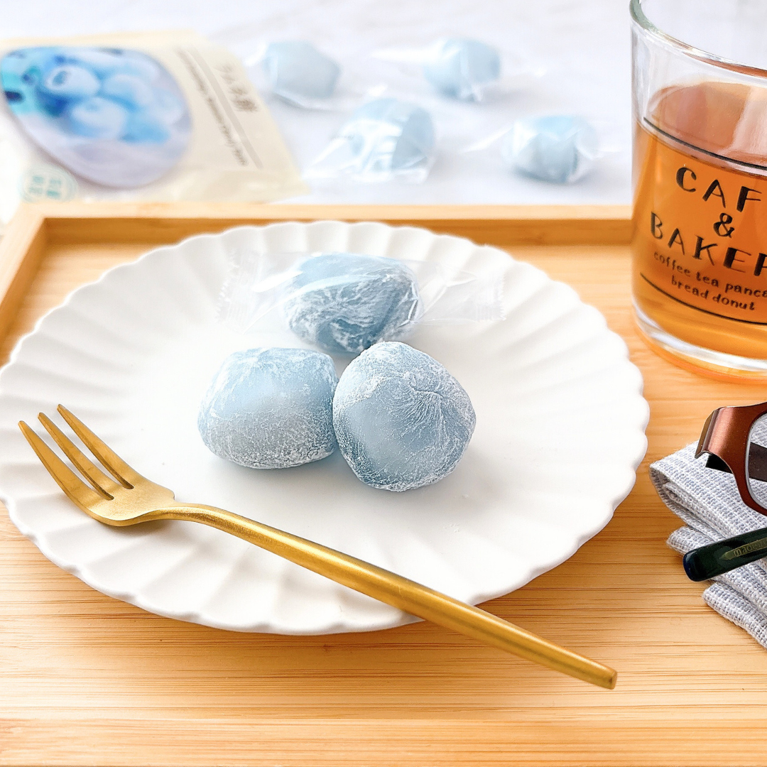 白い皿に青いもち菓子が3つ乗っており、横に金色のフォークがあります。隣には飲み物が入ったグラスがあります。