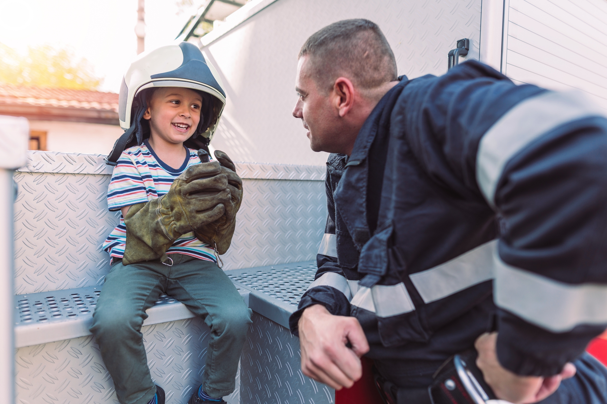 Young child wearing oversized firefighter helmet and gloves sits on fire truck, talking to a smiling firefighter in uniform kneeling beside them