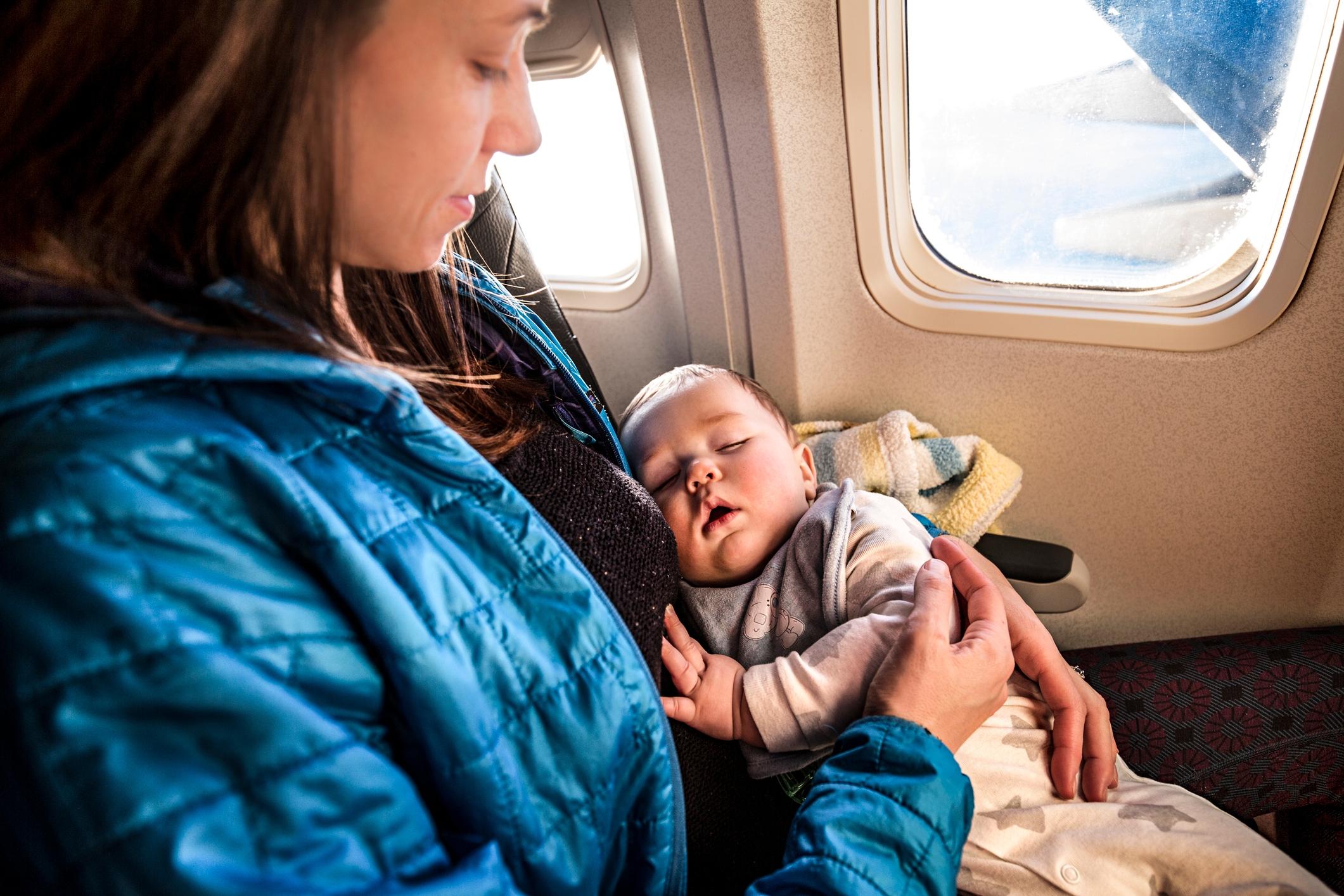 A person on a plane holds a sleeping baby on their lap, next to a window