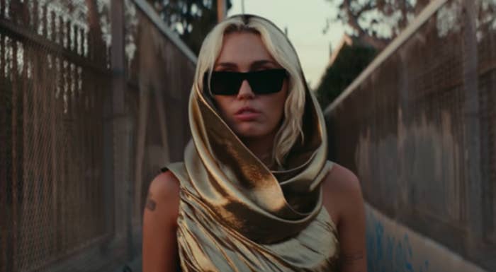 Person in a shiny, draped outfit with a hood and sunglasses, standing on an outdoor walkway