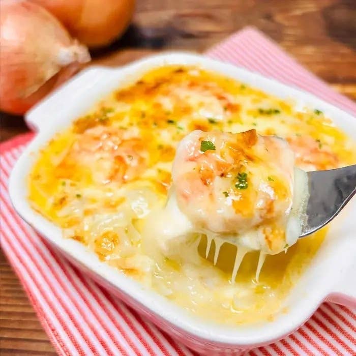 グラタンが白い皿に盛られ、フォークで具材を持ち上げている。背景にタマネギが置かれている。