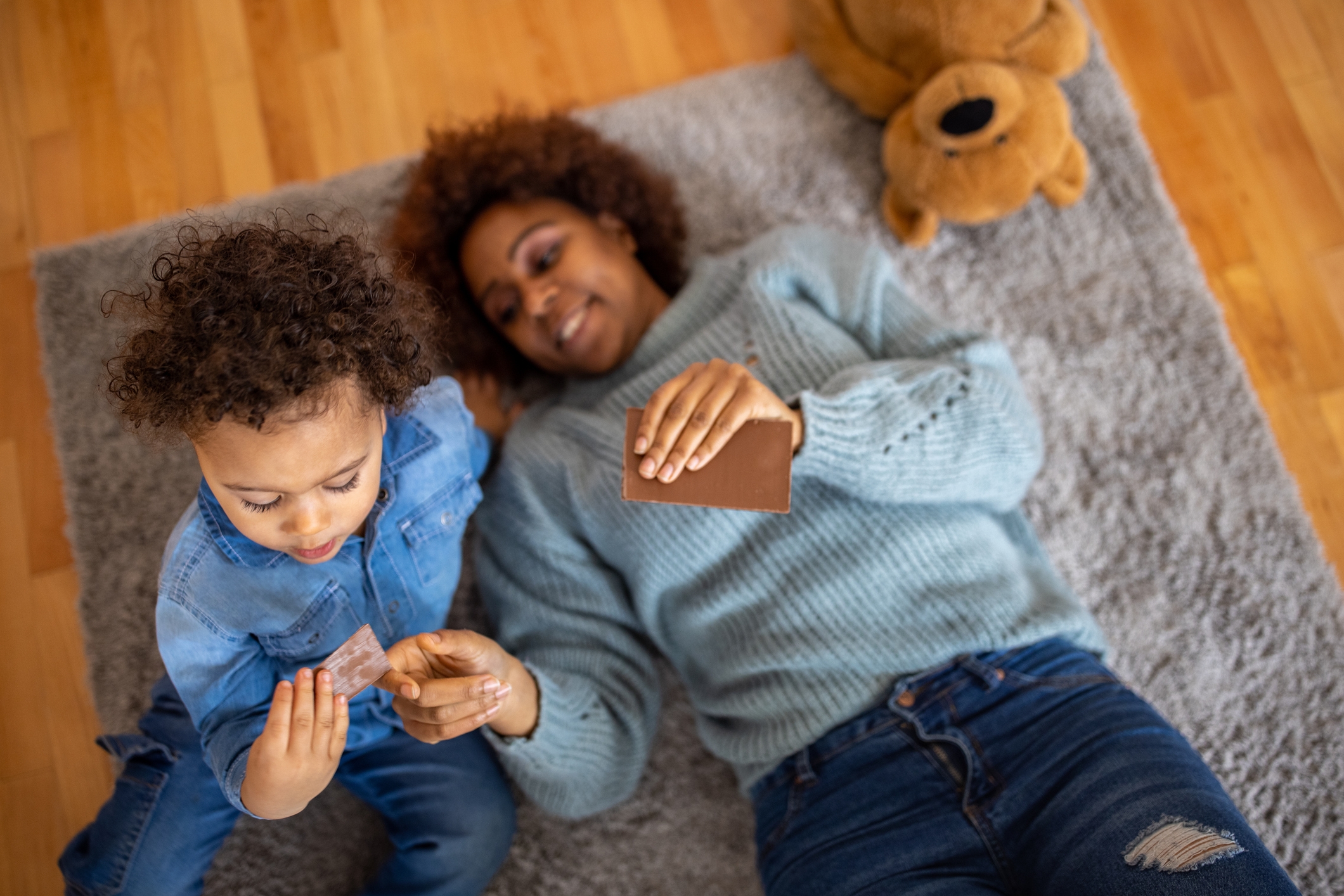 Woman and child lying on a rug, smiling, each holding small photos. A stuffed bear is nearby