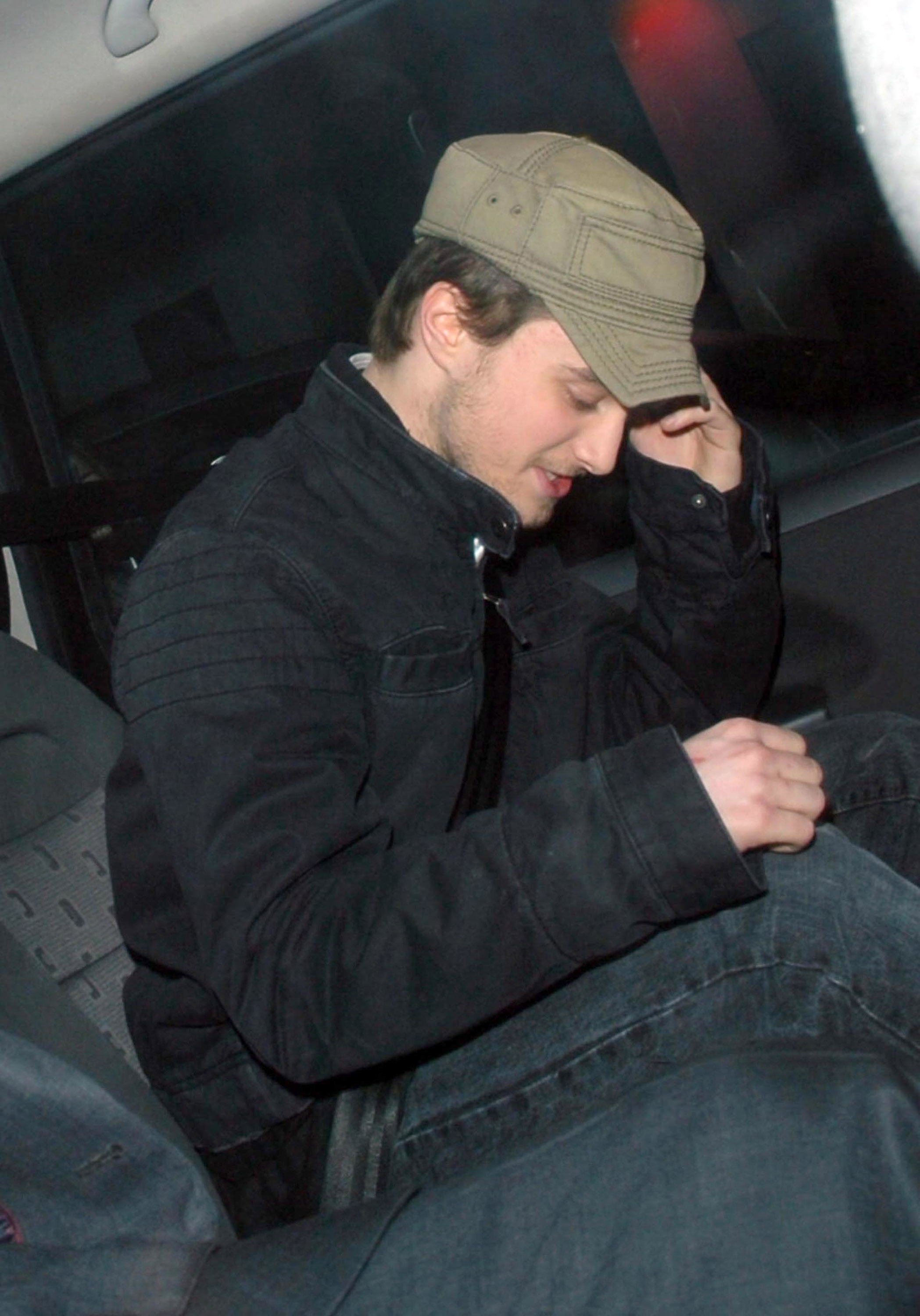Person in a car wearing a casual jacket and cap, looking down while adjusting the cap, seated beside another person whose face is not visible