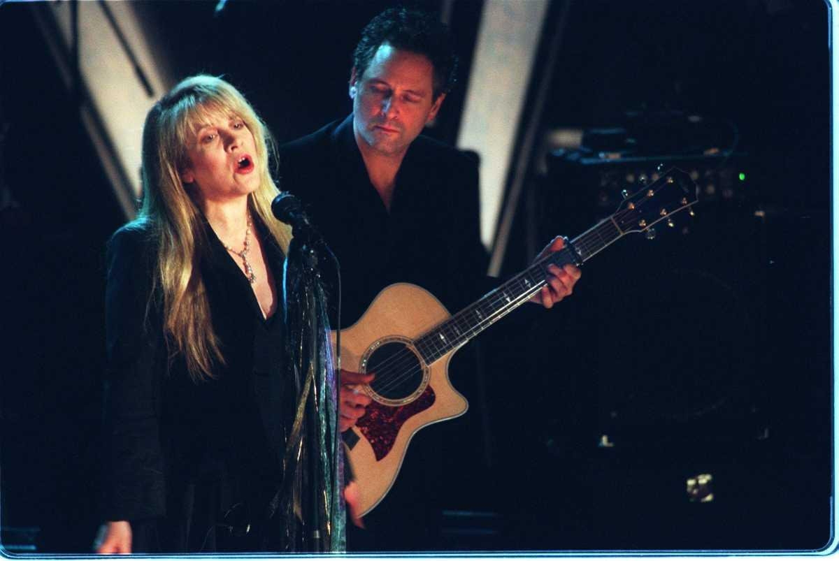A woman sings into a microphone on stage while a man plays guitar behind her. Both are dressed in elegant black outfits
