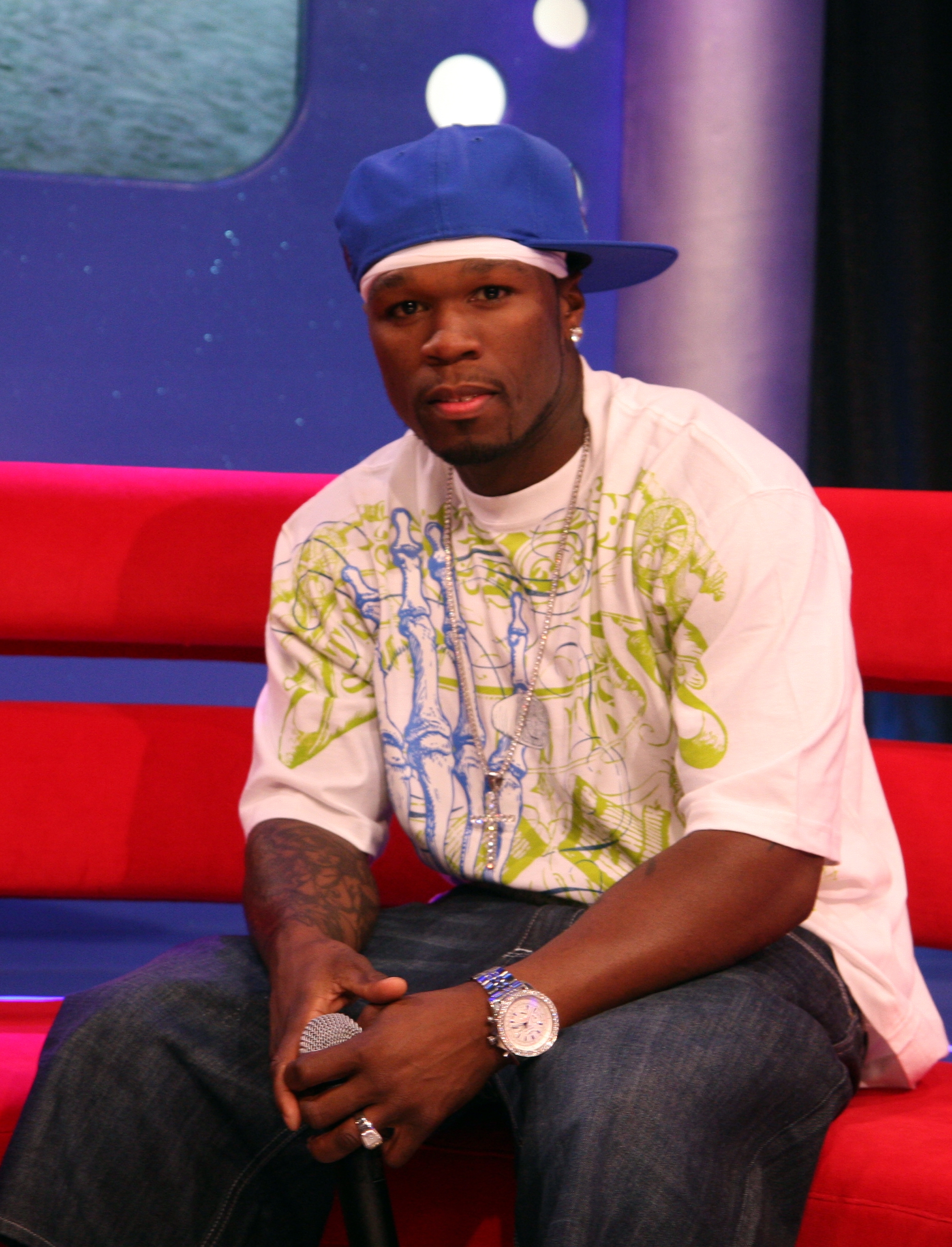 Person sitting on a red couch, wearing a white graphic t-shirt, blue cap, and jeans, holding a microphone