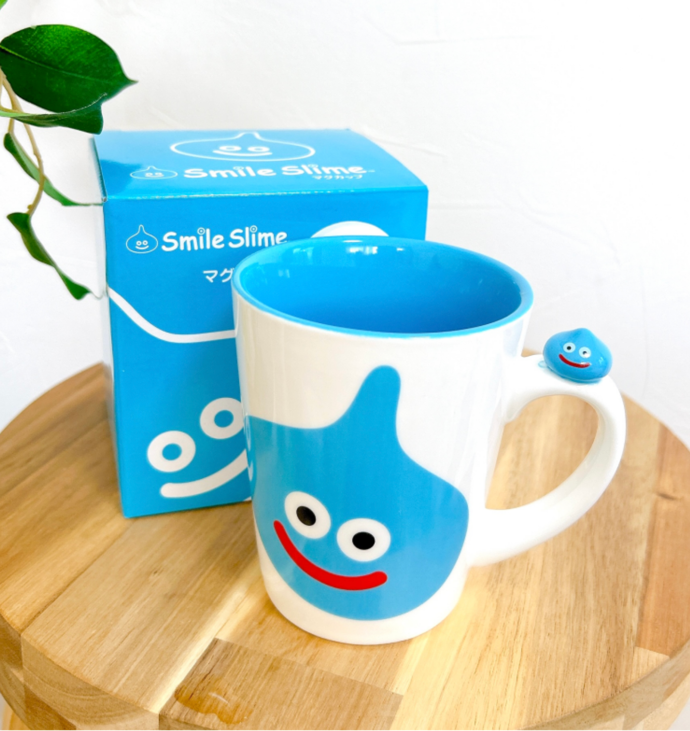 スライムのキャラクターが描かれたマグカップと箱が木のテーブルに置かれている。背景に植物の葉が少し見える。