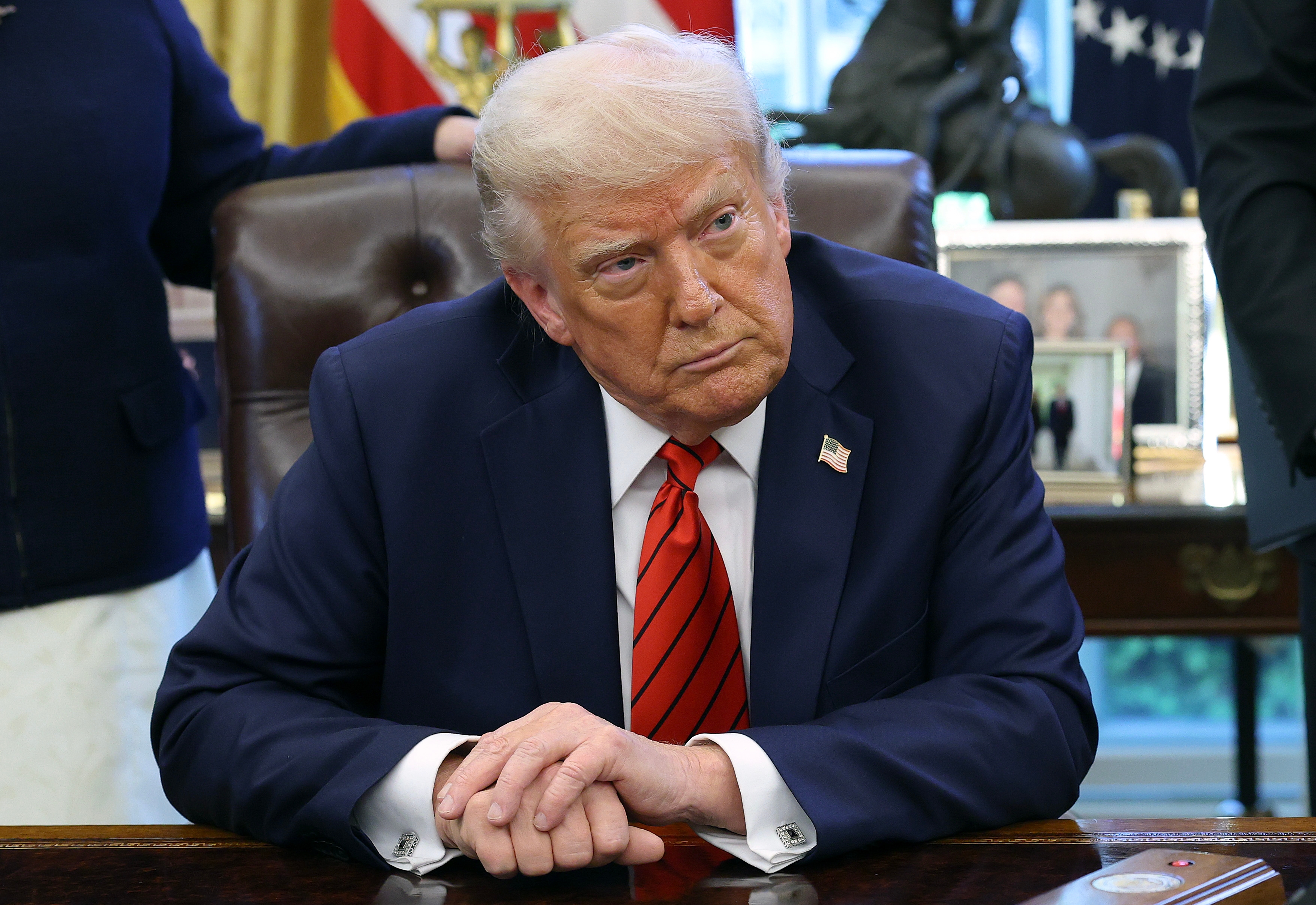 男性がオフィスの机に座る。スーツを着用し、赤いネクタイを締めている。背景に写真や旗が飾られている。