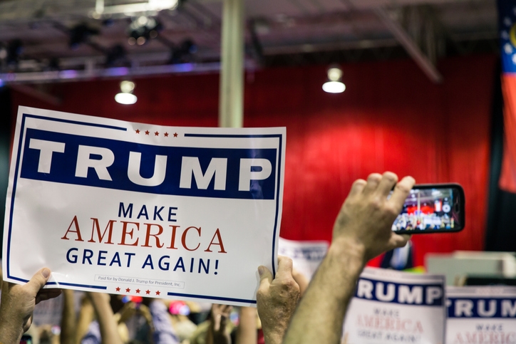 People holding "Trump Make America Great Again" signs at a rally, with a person taking a photo on a smartphone