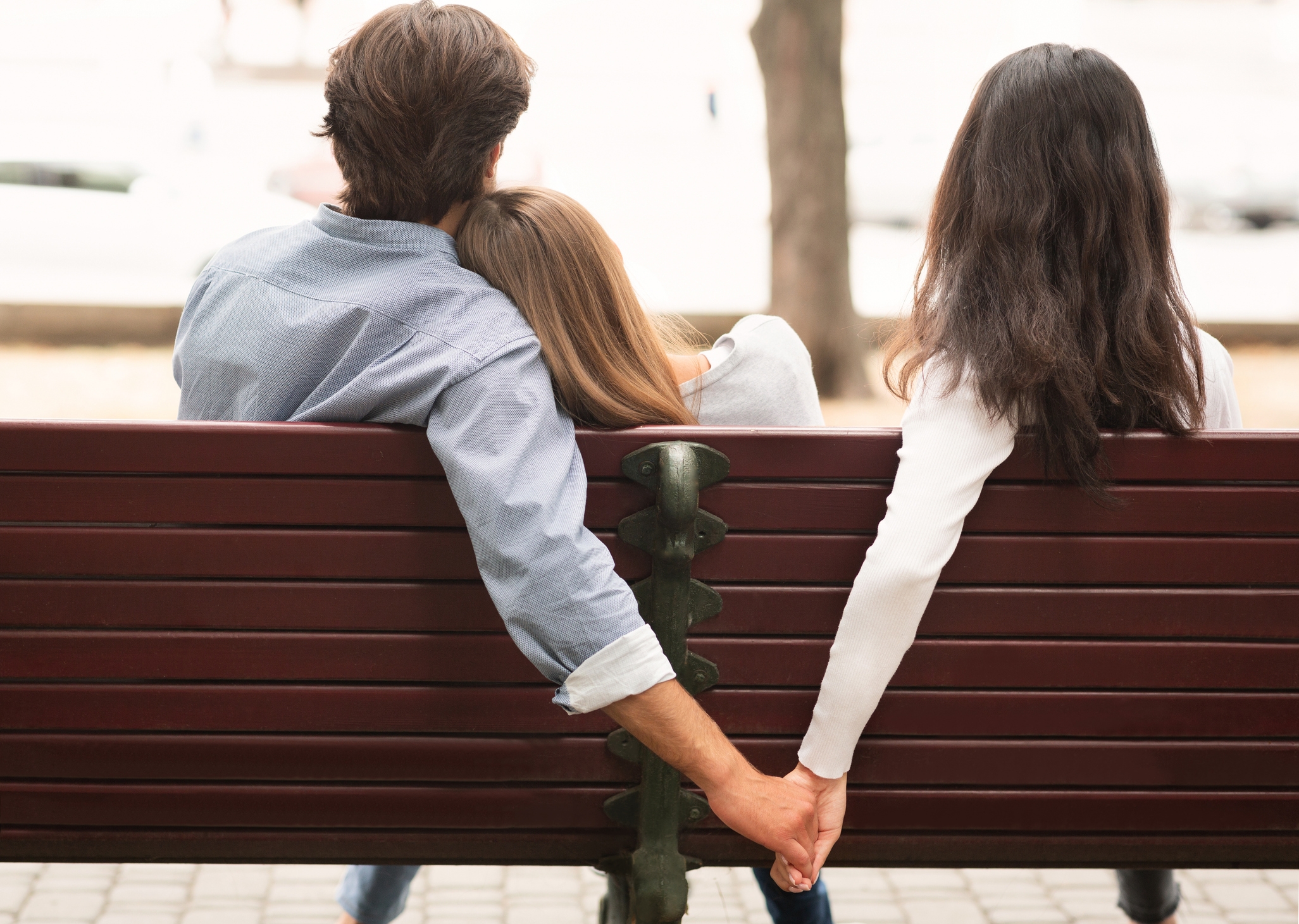 Three people sit on a bench; one person is secretly holding another's hand behind the other's back