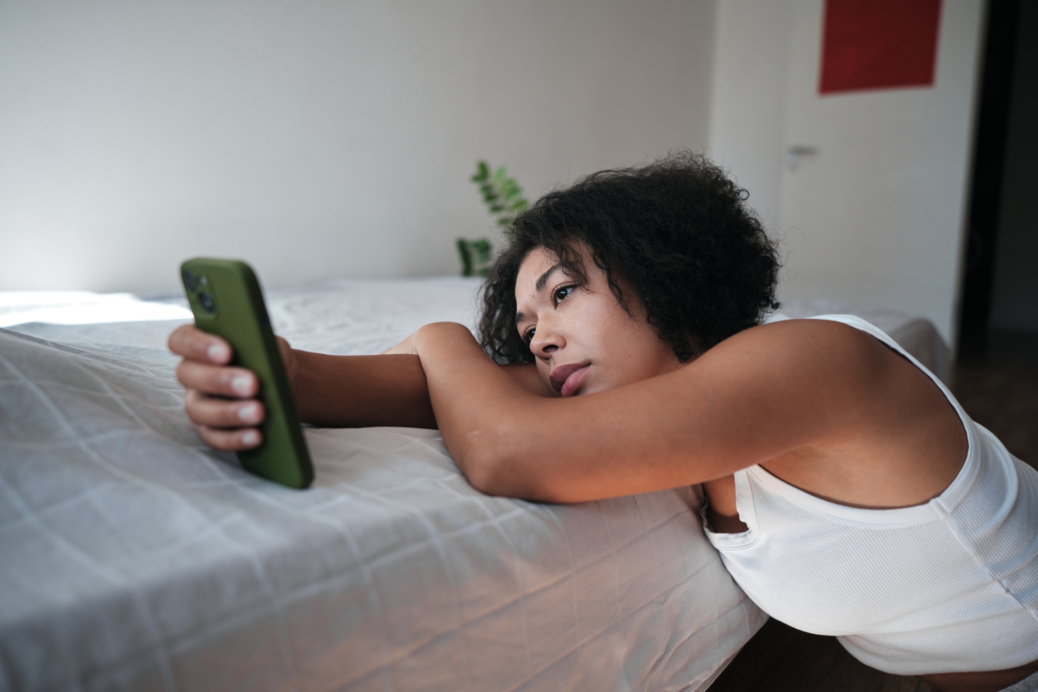 Person in a tank top lies on a bed, looking at their phone with a tired expression
